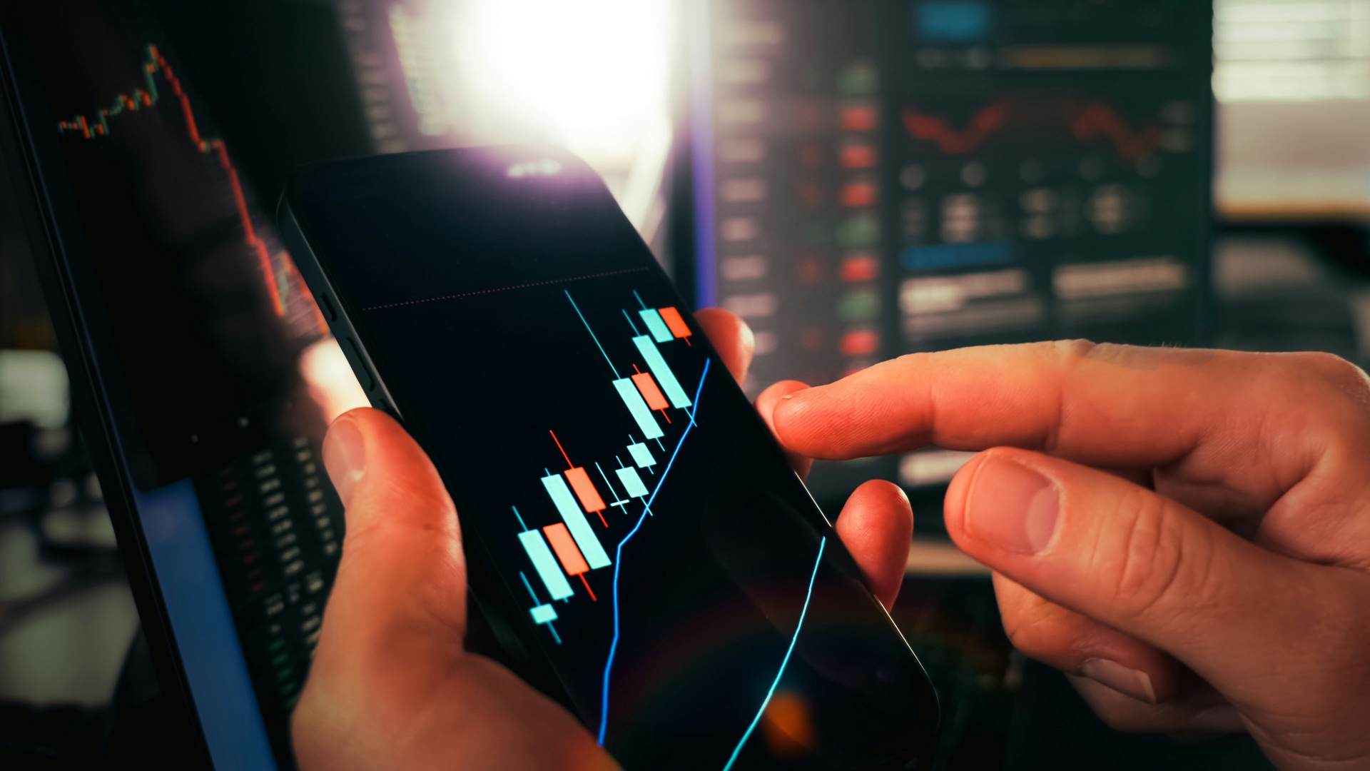 Hands holding a smartphone displaying a candlestick chart with trading screens in the background